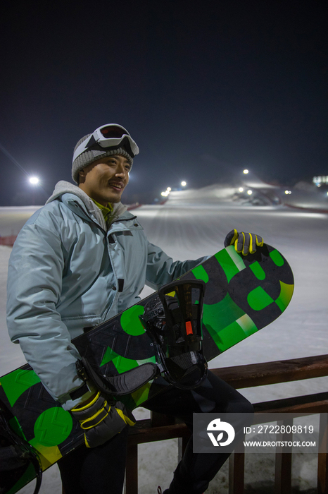 夜晚青年男人抱着滑雪板坐在滑雪场内的护栏上