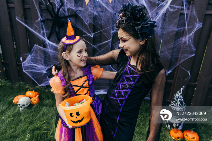 Smiling girls with halloween makeup holding candies in bucket while standing near decor in backyard