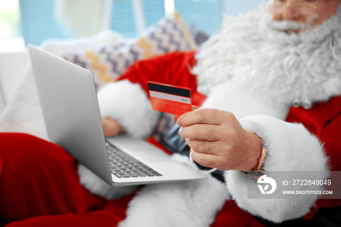 Santa Claus with credit card and laptop on couch at home