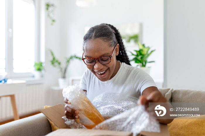 Smiling African woman sit on couch at home open post package shopping online buying goods on interne