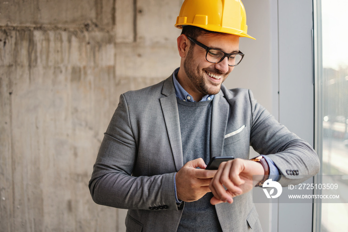 Smiling businessman with helmet standing in building in construction process next to a window, looki