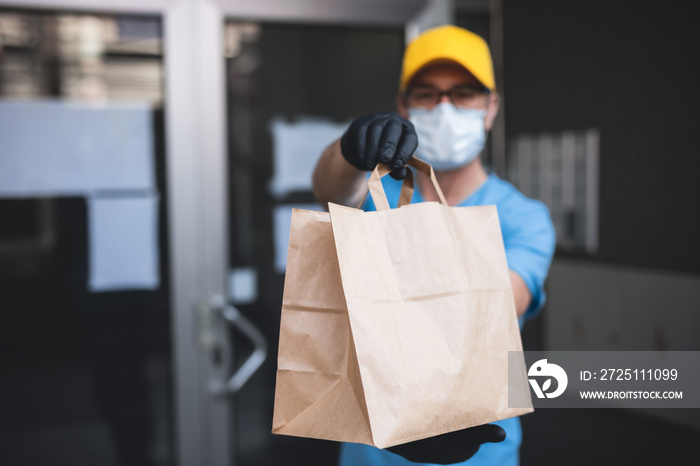 Delivery guy with protective mask and gloves holding box / bag with groceries in front of a building