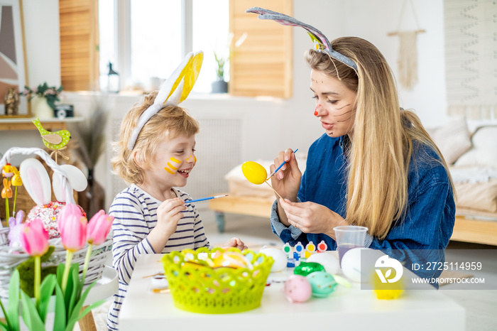 Beautiful happy son and mother in headbands with bunny ears decorate eggs with a brush and bright pa