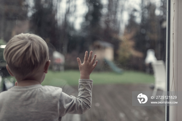 Toddler boy staring out of a window in evening light into the garden