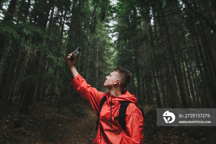 Young backpacker guy in a bright red raincoat taking selfie on his smartphone in the mountain woods.