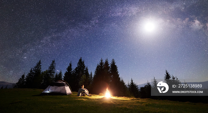男性徒步旅行者独自坐在草地山谷篝火旁的旅游帐篷旁，享受夜晚蓝色的星空