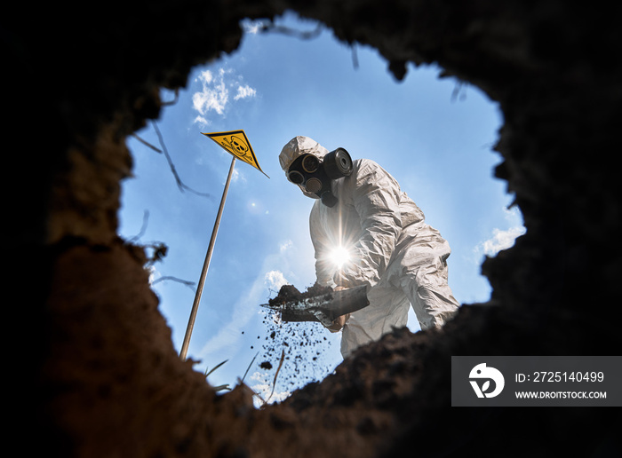 View from inside pit of ecologist digging pit by shovel, wearing gas mask and protective overalls ou