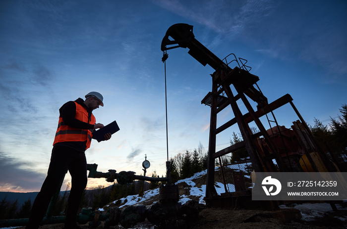 Oil engineer in work vest at oil pump rocker-machine, making notes while checking work of balanced b