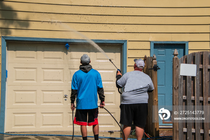 Obese Caucasian man is spray pressure washing the siding on his house. His son is holding the hose a