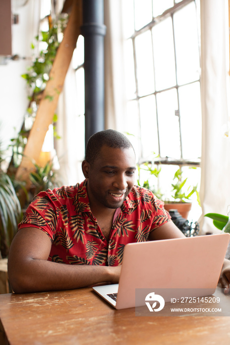 Smiling man using laptop computer on wooden table while sitting by window at home