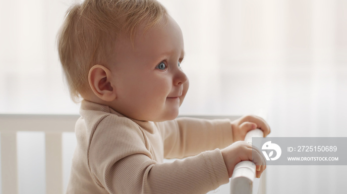 Portrait of adorable little baby standing in crib and smiling aside, looking with curiosity, panoram