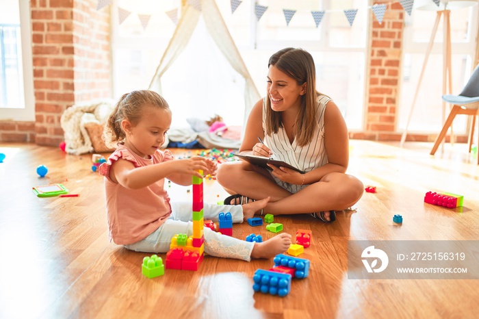 Beautiful psycologist and blond toddler girl doing therapy building tower using plastic blocks at ki