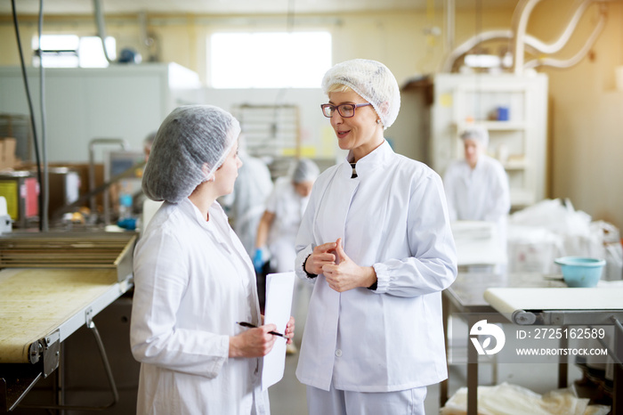 Two young joyful female workers in wearing sterile cloths are discussing statistics of food industry