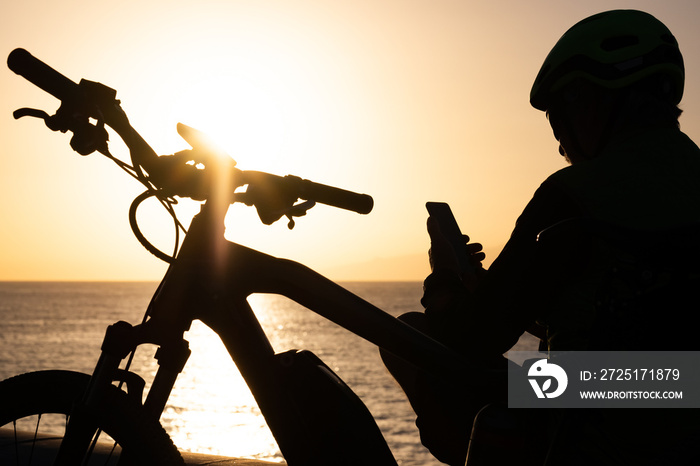 Cyclist man wearing sport helmet sitting on the beach at sunset using mobile phone. Adult mature man