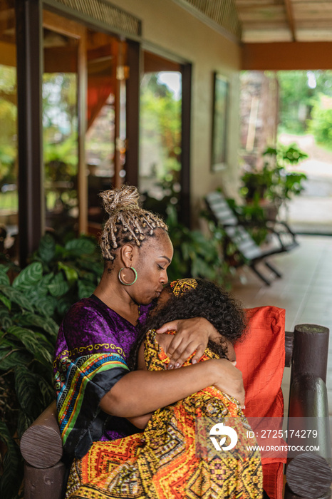 Imagen vertical de una madre afrocaribeña sentada cargando a su hija con mucho amor mientras la besa