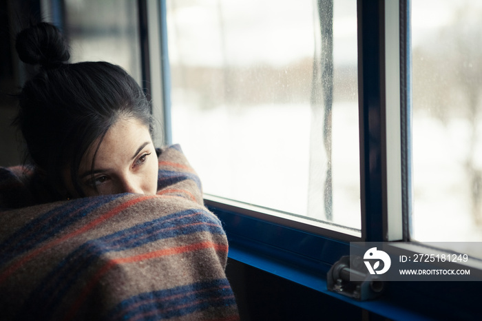 Thoughtful woman wrapped in blanket looking through window while sitting at home during winter