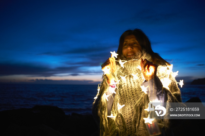 Mature woman wrapped in shawl with star fairy lights on beach at night