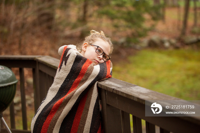 Side view of thoughtful cute girl wrapped in blanket leaning on wooden railing during winter