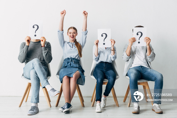 Smiling woman showing yes gesture near employees holding cards with question marks in office