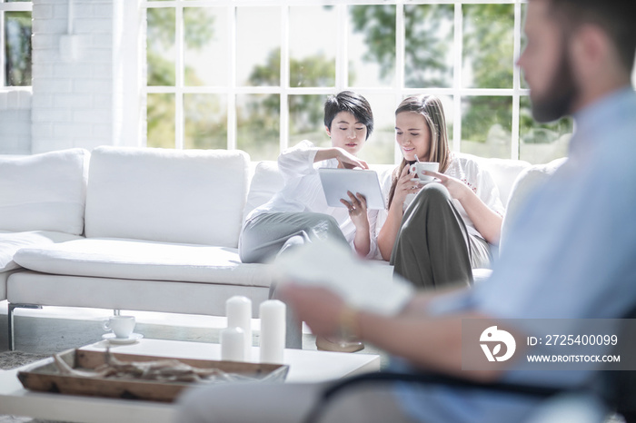 Women using digital tablet on sofa, man reading in foreground