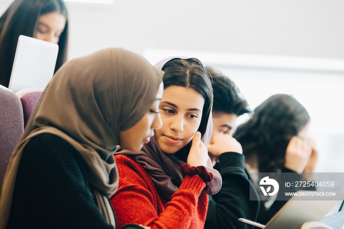 Friends in hijab talking while using laptop in lecture hall