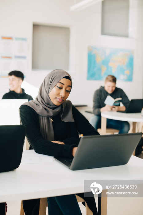 Teenage student wearing hijab using laptop at desk in university classroom