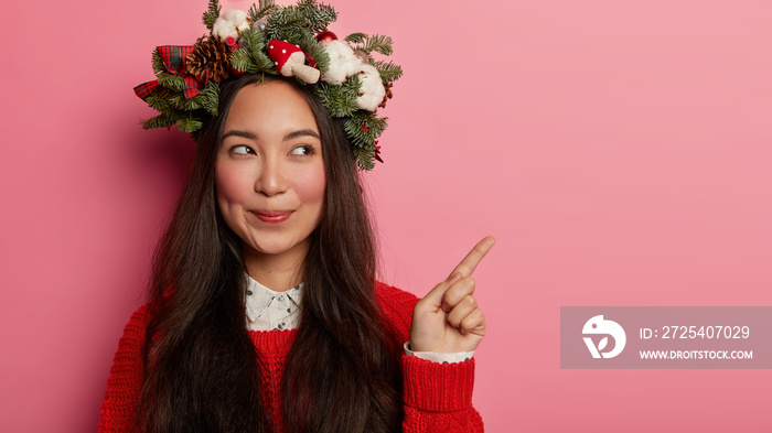 Horizontal shot of attractive brunette woman wears holiday wreath, points fore finger on right side,