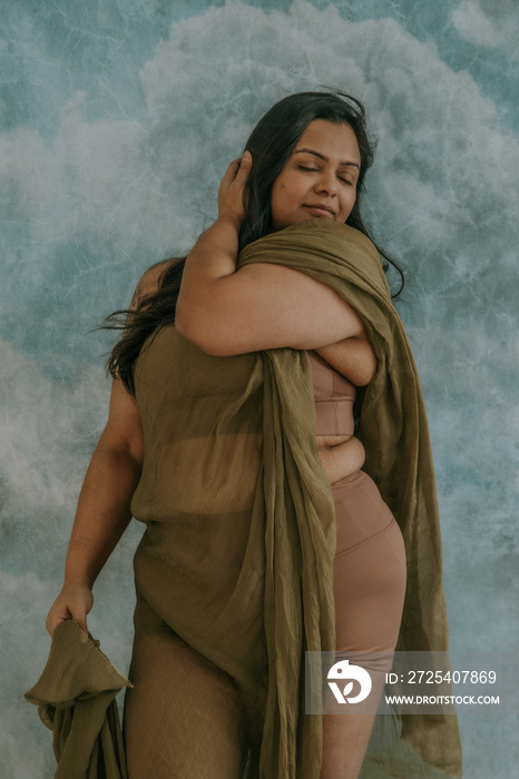 East Indian woman standing with hand in hair wearing green fabric and tan bra and short eyes closed