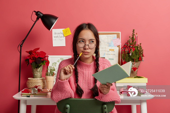 Serious contemplating Asian girl with combed plaits, holds notebook and pencil, writes down future p