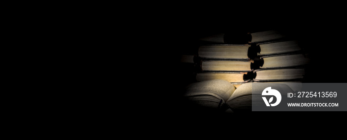 aufgeschlagenes Buch in düsterem Licht vor einem Bücherstapel mit viel dunklem Textfreiraum