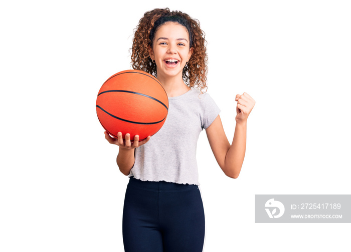 Beautiful kid girl with curly hair holding basketball ball screaming proud, celebrating victory and 