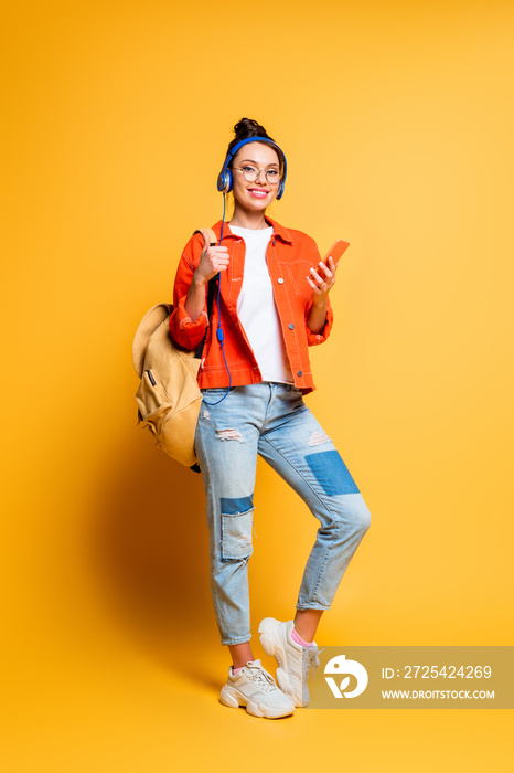 attractive smiling student in headphones holding backpack and smartphone while looking at camera on 