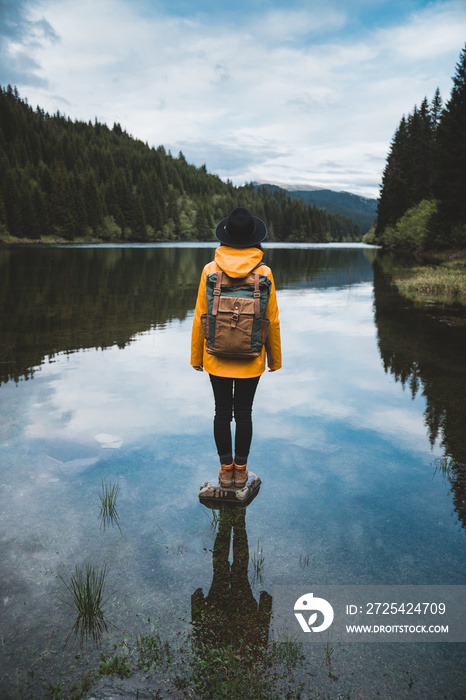 Back view of stylish hipster woman wearing backpack, hat and yellow jacket looking at mountain view 