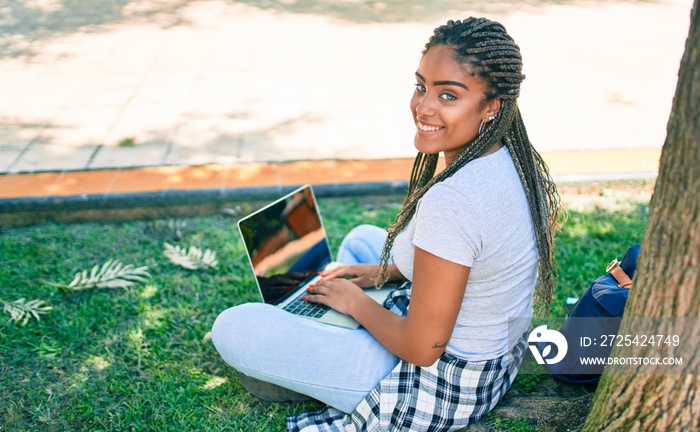 Young african american student woman smiling happy using computer laptop sitting on the grass at the