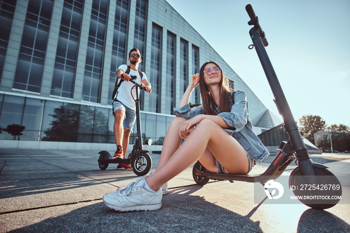 On a bright sunshining day couple of young people chilling near big building with their scooters. Lo