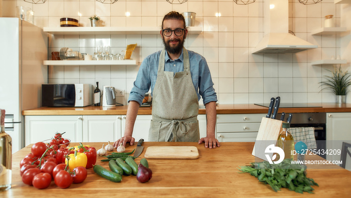 Young man, Italian cook in apron smiling at camera while preparing healthy meal with vegetables in t