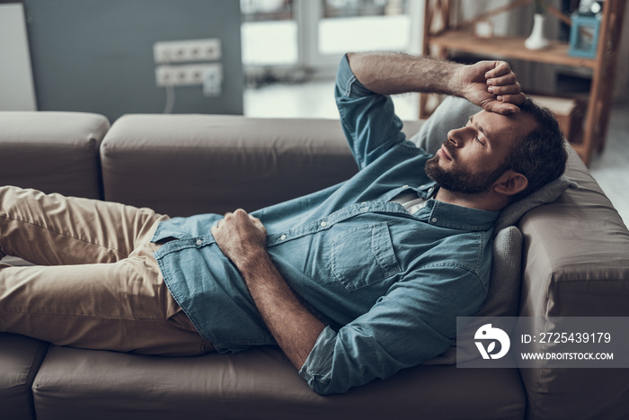 Adult man lying on the sofa and closing eyes while being at home