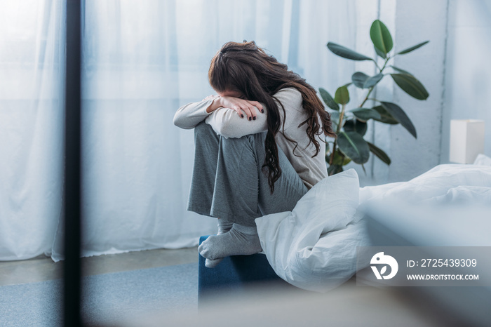 grieving woman sitting, covering face with hands and crying in bedroom