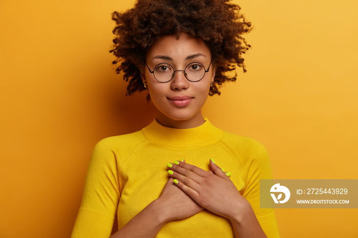 Serious calm Afro woman presses hands to heart in grateful gesture, appreciates nice words, expresse