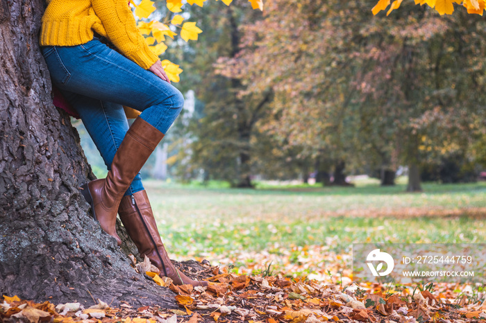 时尚女士在秋季公园靠在树上。穿着牛仔裤和皮靴的女士腿。