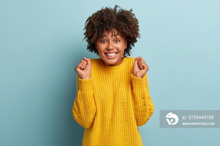 Portrait of cheering dark skinned female with curly hair clenches fists in victory gesture, happy to