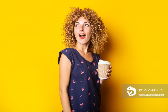 surprised young woman with curly hair holding paper cup on yellow background.