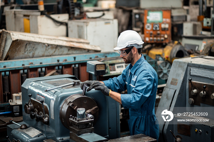 Men industrial engineer wearing a white helmet while standing in a heavy industrial factory behind. 