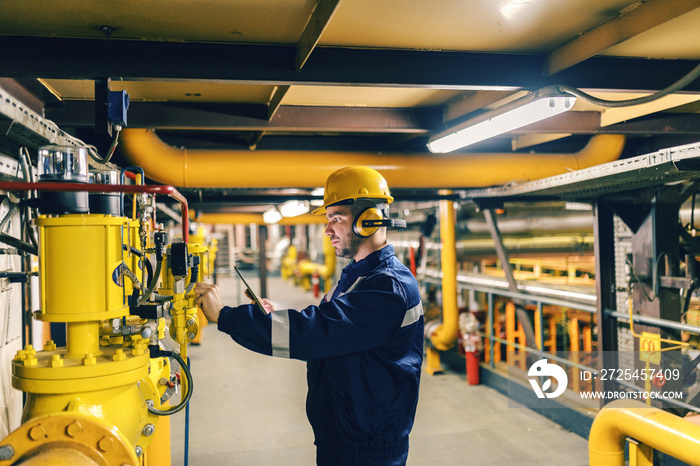 Young Caucasian worker in protective suit using tablet while checking machines in heating plant.