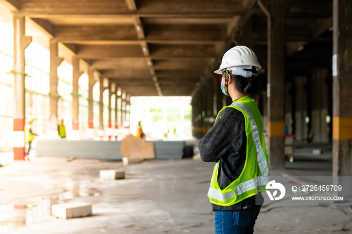 Behind a woman, a construction engineer wearing a hat and a green safety vest, stands looking at wor