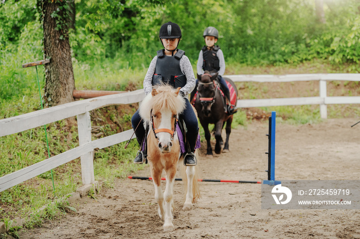 Children with helmets and protective vests on riding pony horses at sunny day on ranch.