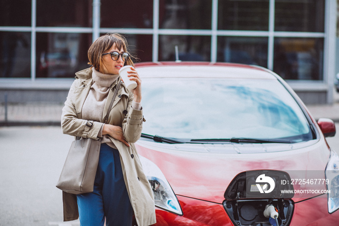 Woman charging electro car at the station and drinking coffee