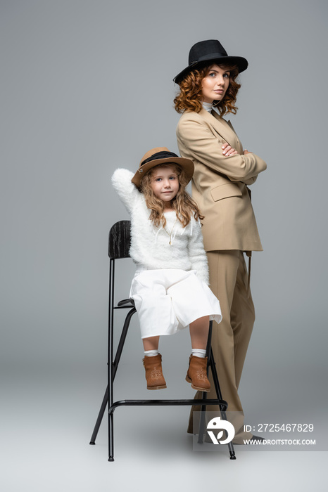 elegant mother and daughter in white and beige outfits and hats posing on chair on grey background