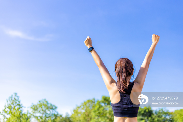 Back view of a woman in stretching up her arms happily at an outdoor field with blurred trees and cl