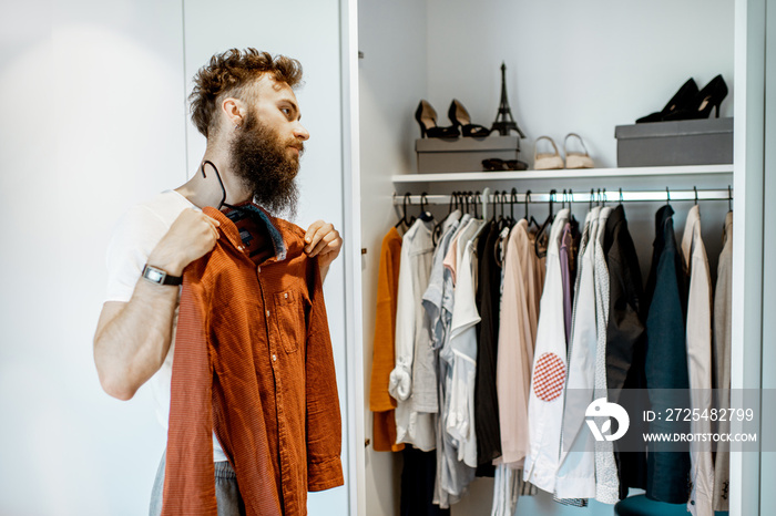 Bearded man trying shirt, choosing clothes to wear in the wardrobe at home